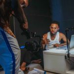 Dois jovens negros gravam um vídeo em um estúdio doméstico, com câmera, laptop e equipamentos de produção ao redor.