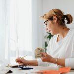 Empreendedora calcula as finanças com dinheiro, calculadora e caderno sobre a mesa.
